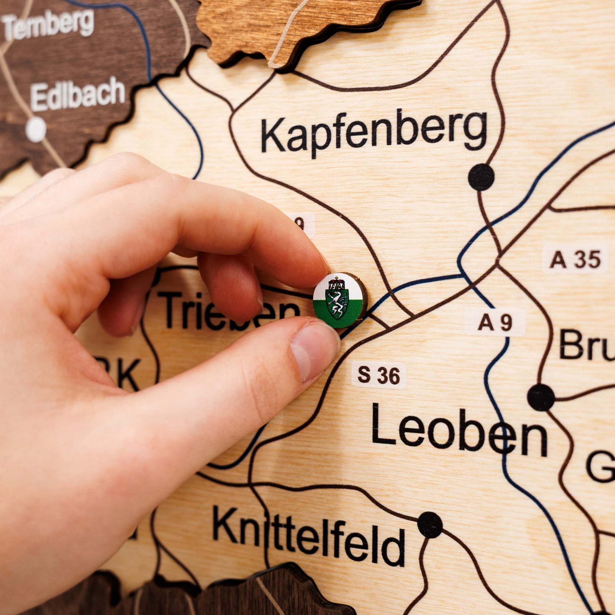Hand placing a badge on a wooden map of Leoben, Kapfenberg, and Knittelfeld.